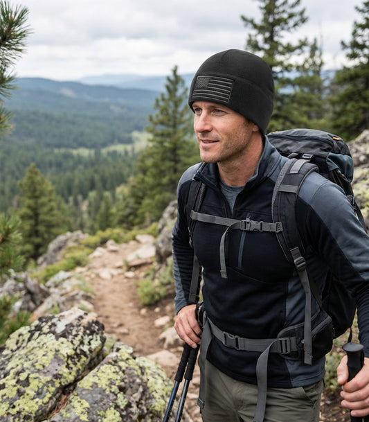 Man hiking with a black beanie with American flag on it in a mountainous area with trees and a clear sky.